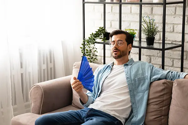 Man sitting on couch in warm house using hand fan